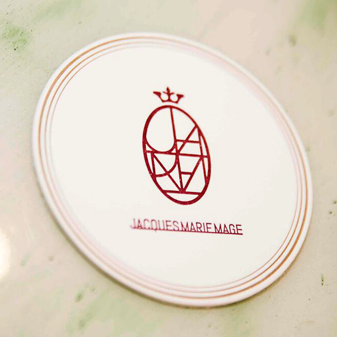 Rundes Emblem in Elfenbein-weiß mit dem roten Logo von Jacques Marie Mage, das sich aus der französischen Bourbonen Lilie ableitet.