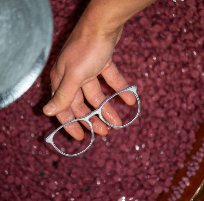 Ein helles Brillenmittelteil auf einer Hand vor rotem Granulat einer Poliermaschine für Brillen aus natürlichem Material, 3D gedruckt.