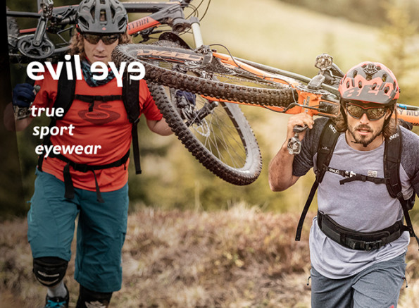 2 Mountainbiker mit ihren Bikes auf dem Rücken und evil eye Sportbrillen laufen einen Berg hoch.