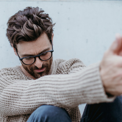 Man in naturfarbenen Wollpullover und schöner Brillenfassung in entspannter Haltung und einem Lächeln im Gesicht.