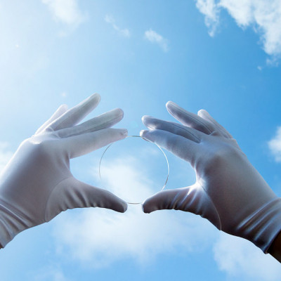 Fast unsichtbares Brillenglas, gehalten von 2 Händen in weißen Handschuhen vor blauem Himmel mit Wölkchen.